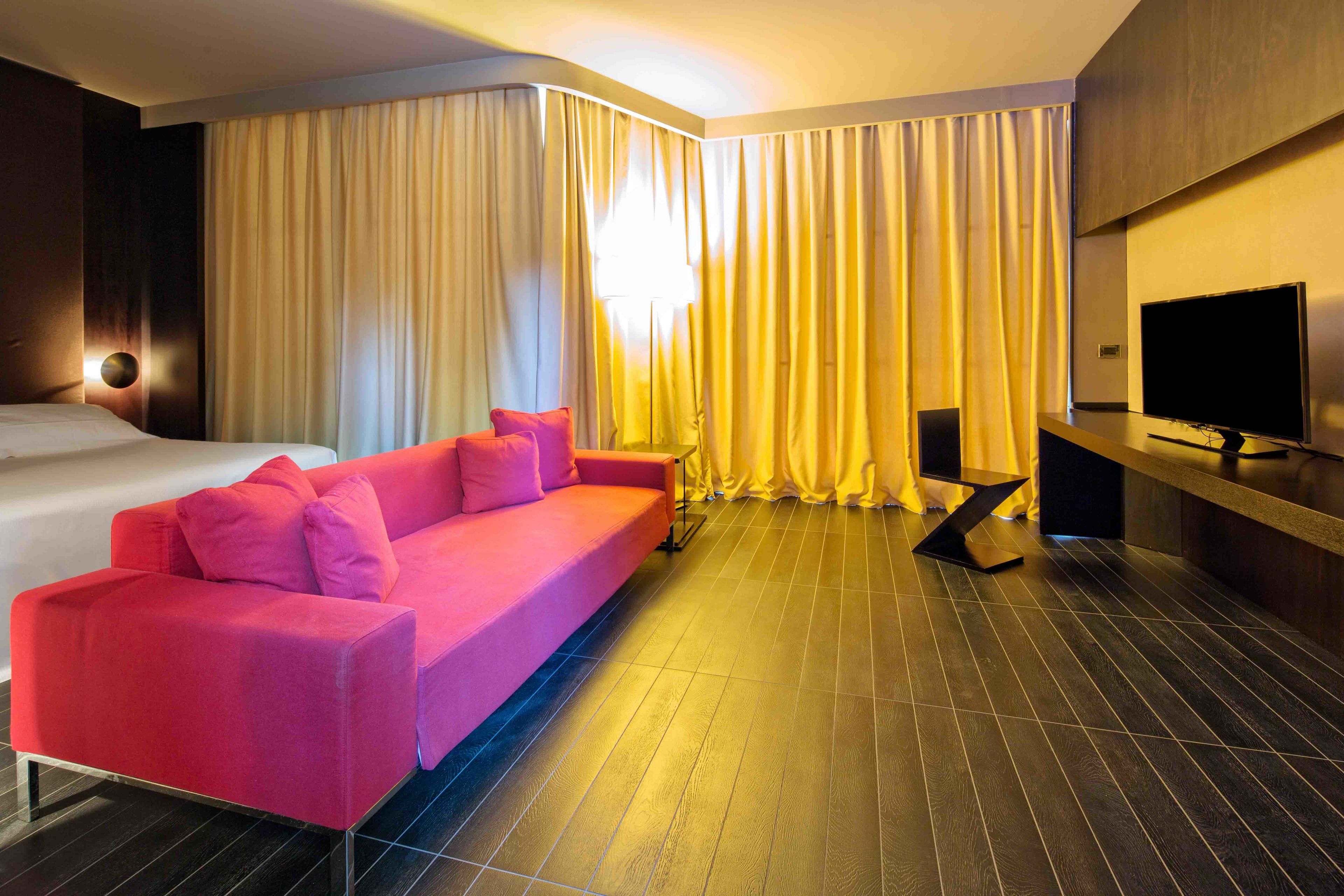 Una habitación de hotel con una cama grande, un sofá rosa, un televisor y cortinas.