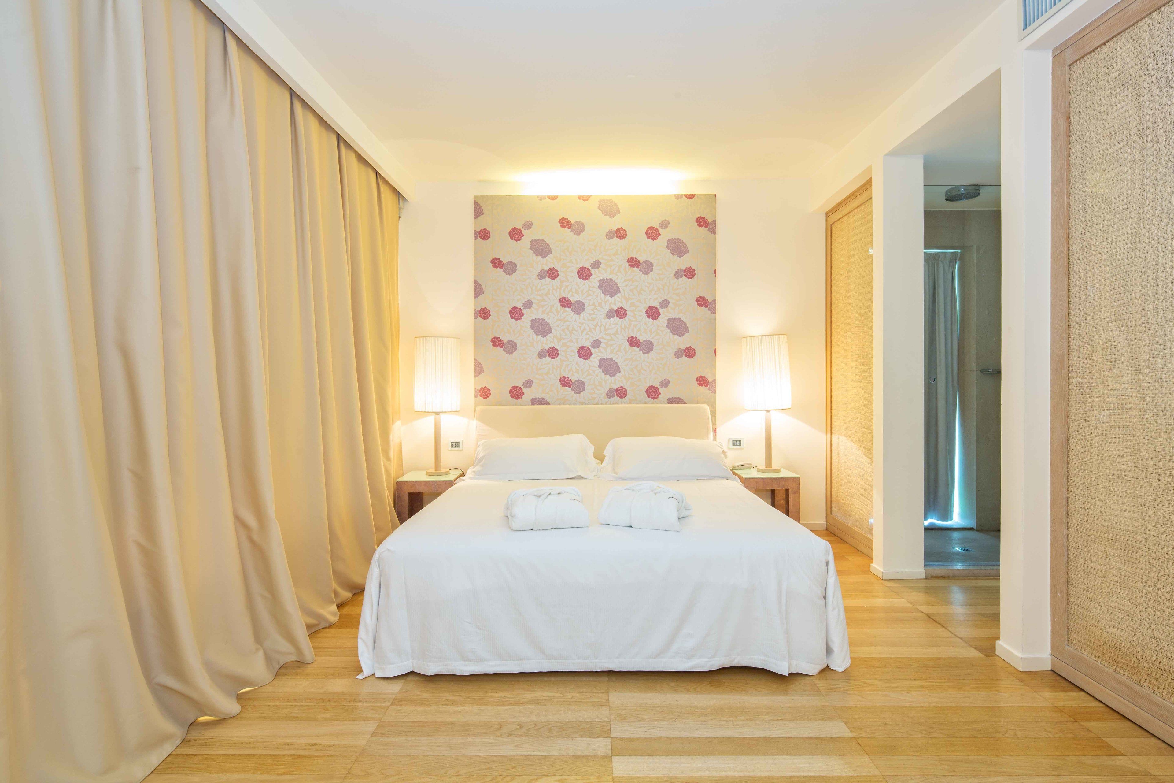 Habitación de hotel con una cama grande, dos lámparas de noche, un papel tapiz estampado con motivos florales y suelo de madera.