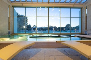 An indoor swimming pool with clear glass walls, two lounge chairs, and a view of the surrounding area.