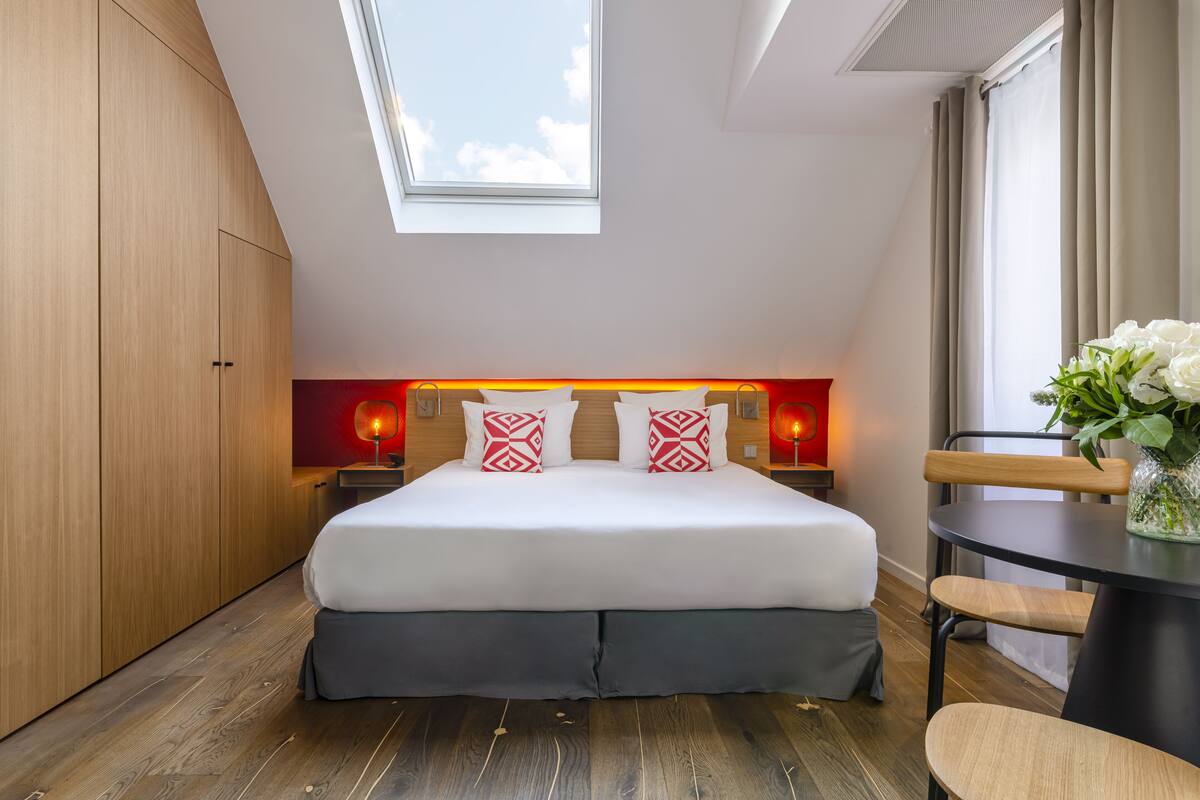 A modern bedroom with a skylight, a large bed with patterned pillows, bedside lamps, a small table with a vase, and a view of the sky.