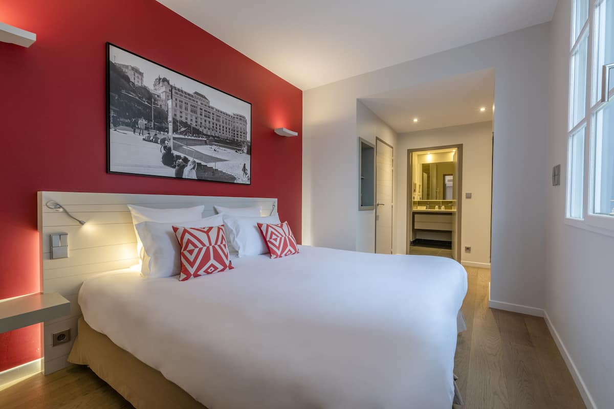 A modern hotel room with a large bed, a red accent wall, and a framed photograph above the bed.