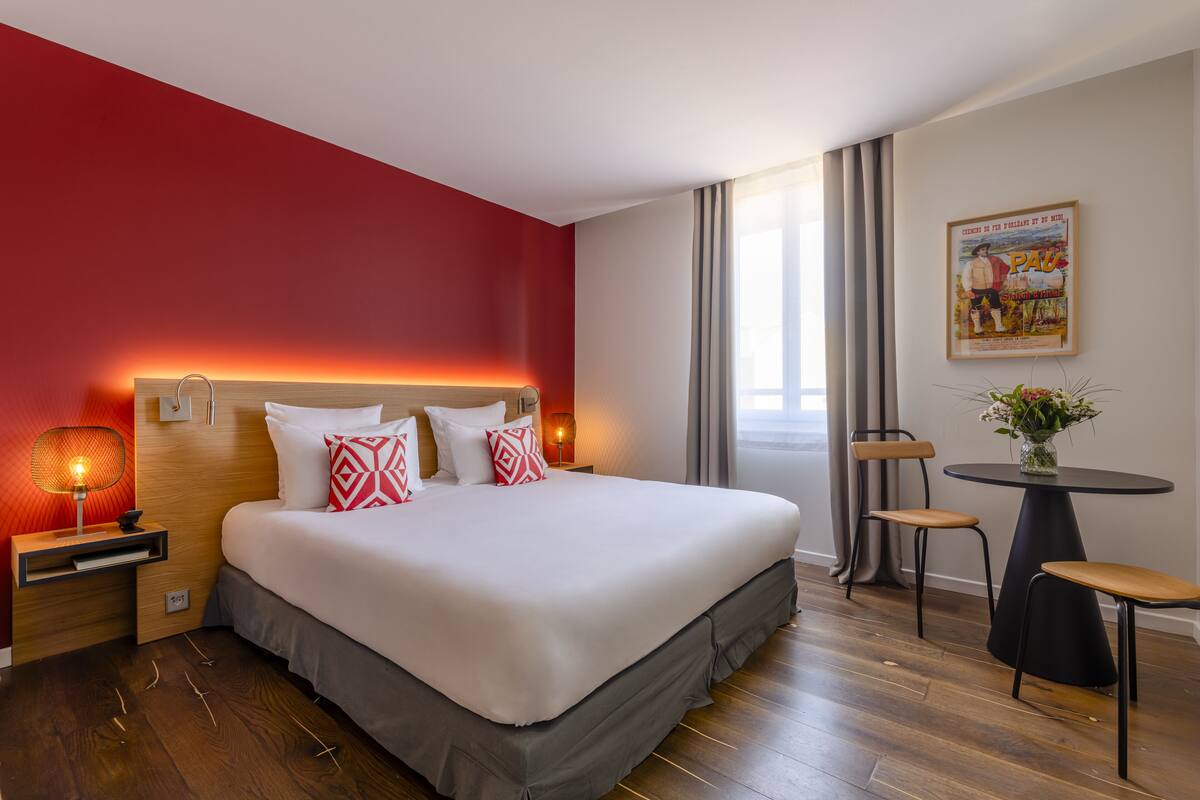 A hotel room with a large bed, a bedside table with a lamp, a small table with two chairs, and a window with curtains.