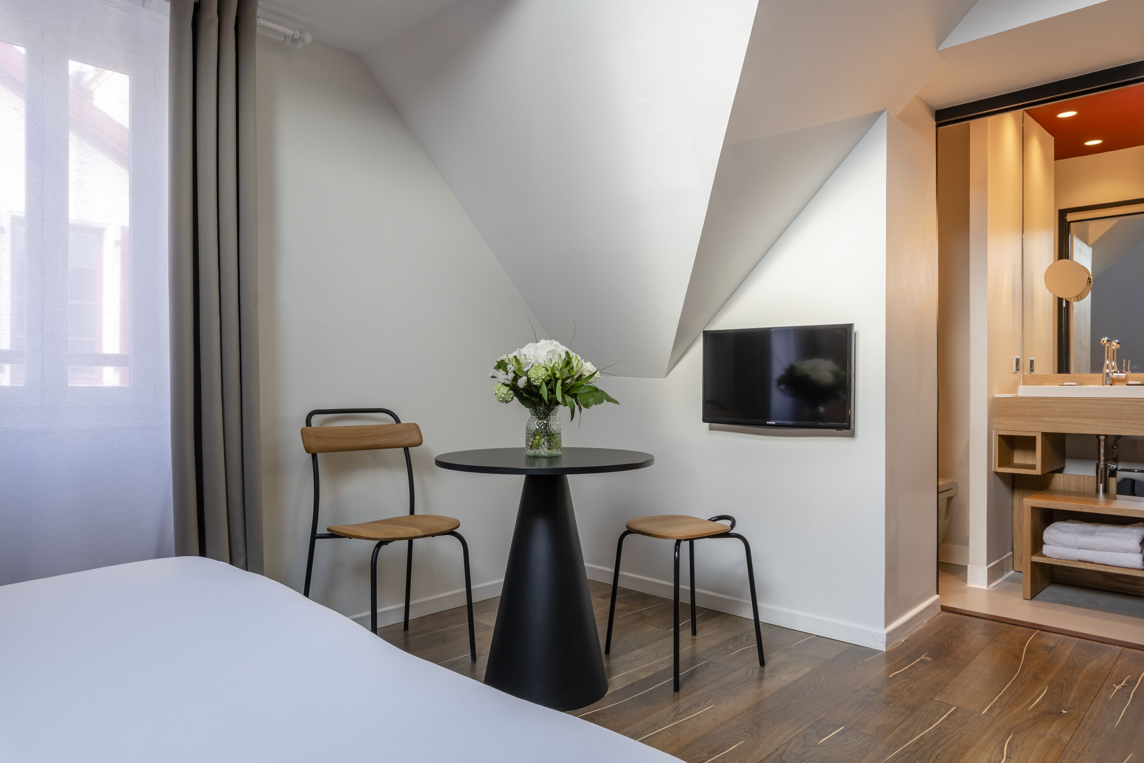 A modern hotel room with a large bed, a round table with chairs, a TV mounted on the wall, and a wooden vanity with a mirror.