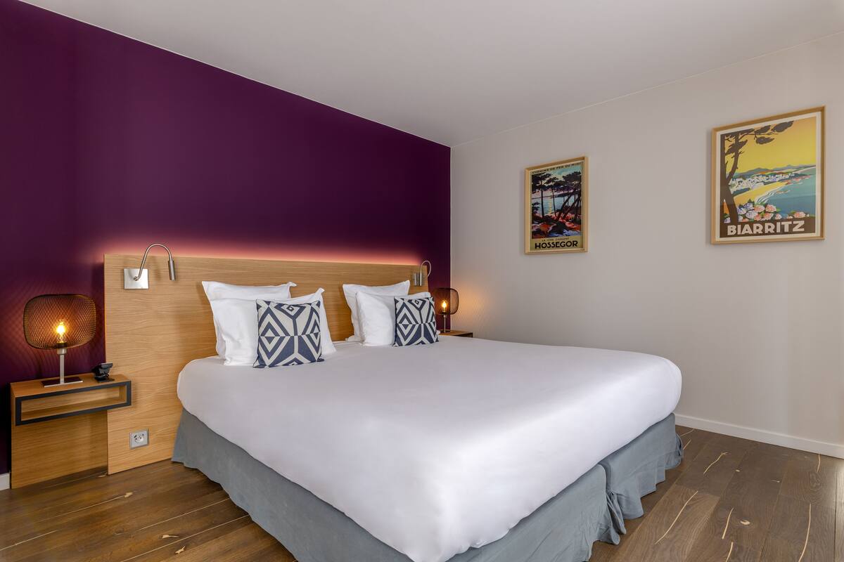 A modern hotel room with a large bed, wooden headboard, and a purple accent wall.