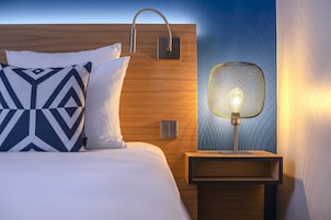 Una cama bien hecha con almohadillas estampadas, una lámpara de pared y una mesita de noche con lámpara.