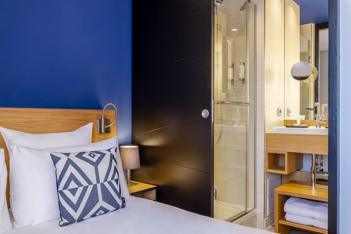 A hotel room with a bed, a nightstand, a bedside lamp, a shower area, and a bathroom with a mirror and sink.