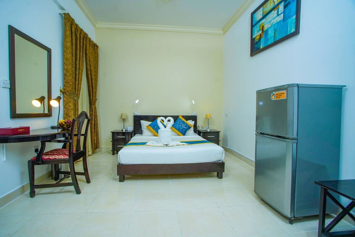 A hotel room with a bed, a desk, a chair, a refrigerator, and a mirror.
