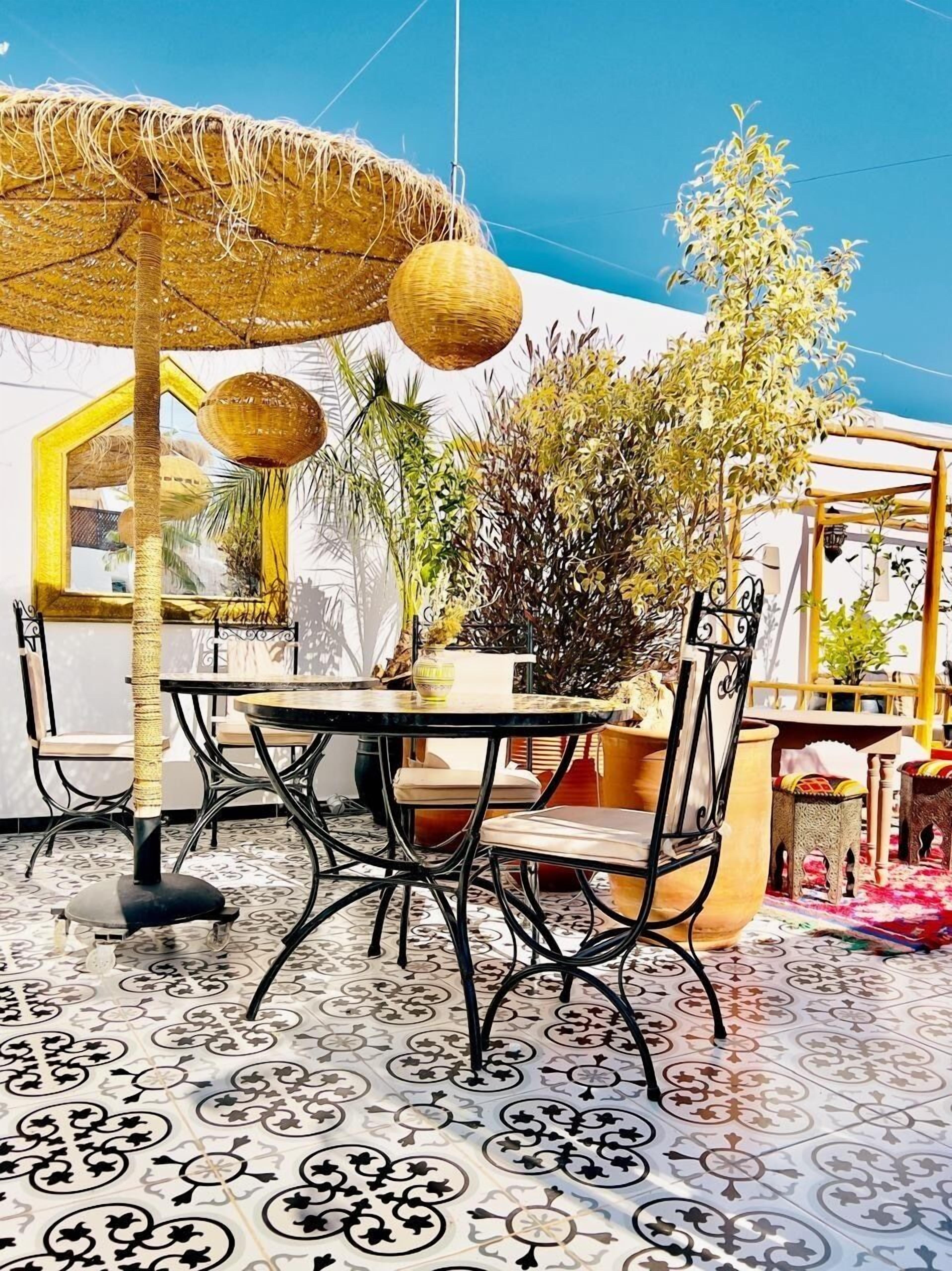 Un área de comedor al aire libre con una mesa redonda, sillas, un paraguas tejido, decoraciones esféricas colgantes y plantas en macetas.