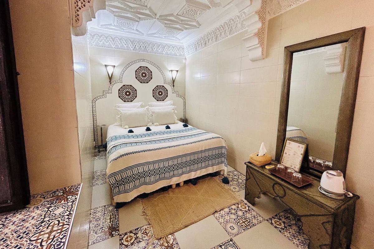 A bedroom with a bed, a nightstand, a mirror, and patterned tiles.