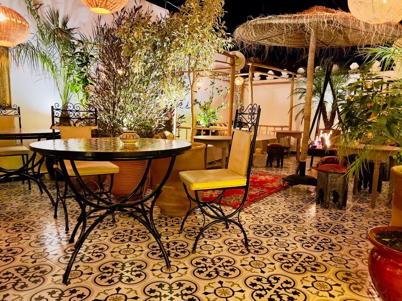 An outdoor dining area with round tables and chairs, surrounded by potted plants and a patterned floor.