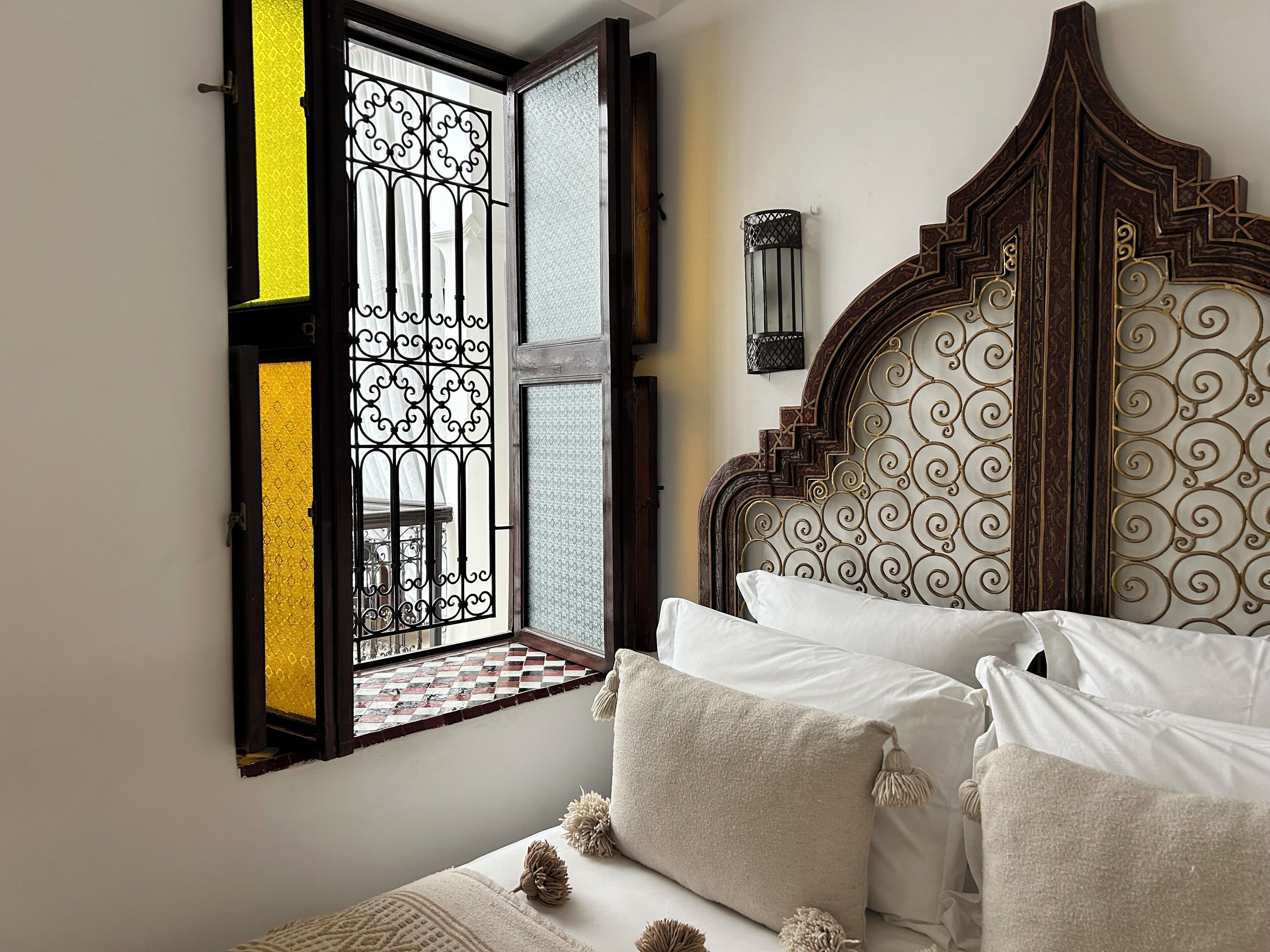 A bedroom with a bed, a decorative headboard, a window with a metal grille, and a wall-mounted light fixture.