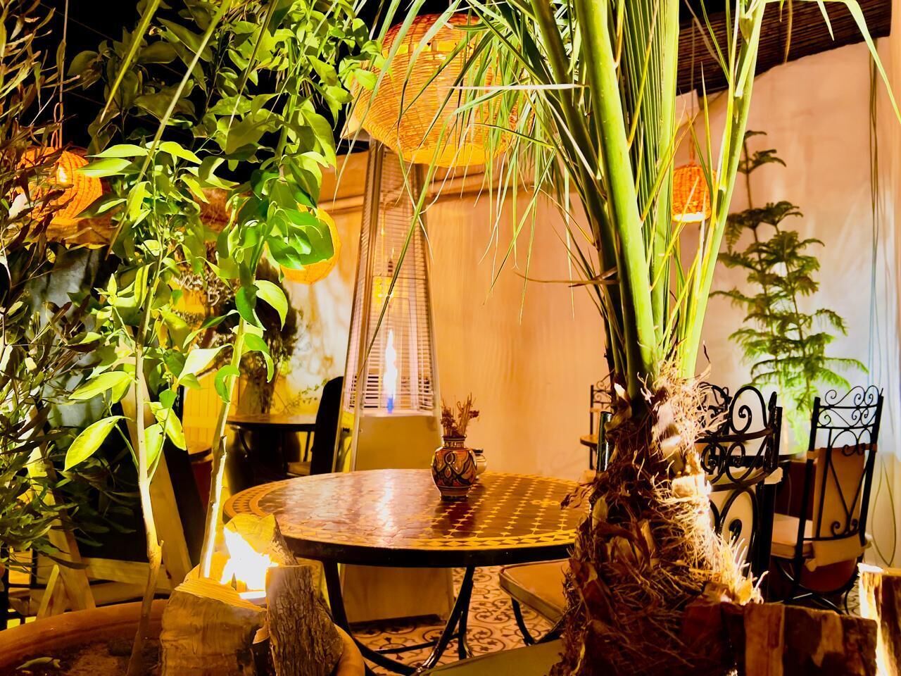 A round table with a woven centerpiece, surrounded by chairs with metal frames, in a room with plants and a window with blinds.