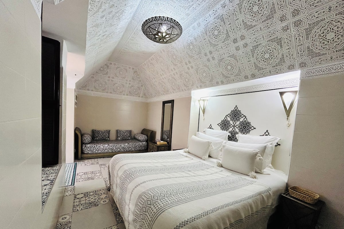 A bedroom with a large bed, a sofa, a mirror, and a decorative ceiling.