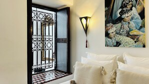 Une chambre à coucher comprenant un lit, des oreillers, une lampe fixée au mur, une fenêtre ornée de barreaux en fer décoratifs et un tableau.