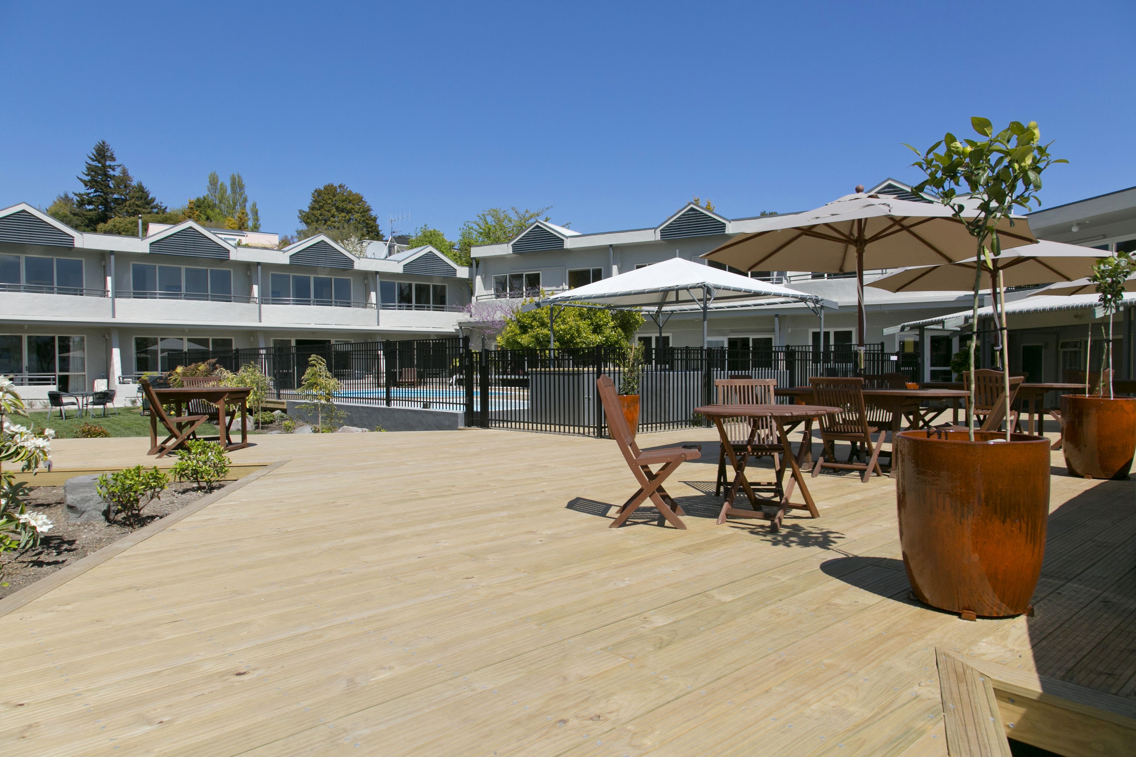 A wooden deck with outdoor furniture and umbrellas, a pool, and a building with multiple windows and balconies.