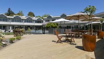 A wooden deck with outdoor furniture and umbrellas, a pool, and a building with multiple windows and balconies.