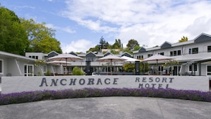Exterior del Anchorage Resort Motel, con un letrero, asientos exteriores con sombrillas y un área ajardinada con flores moradas.