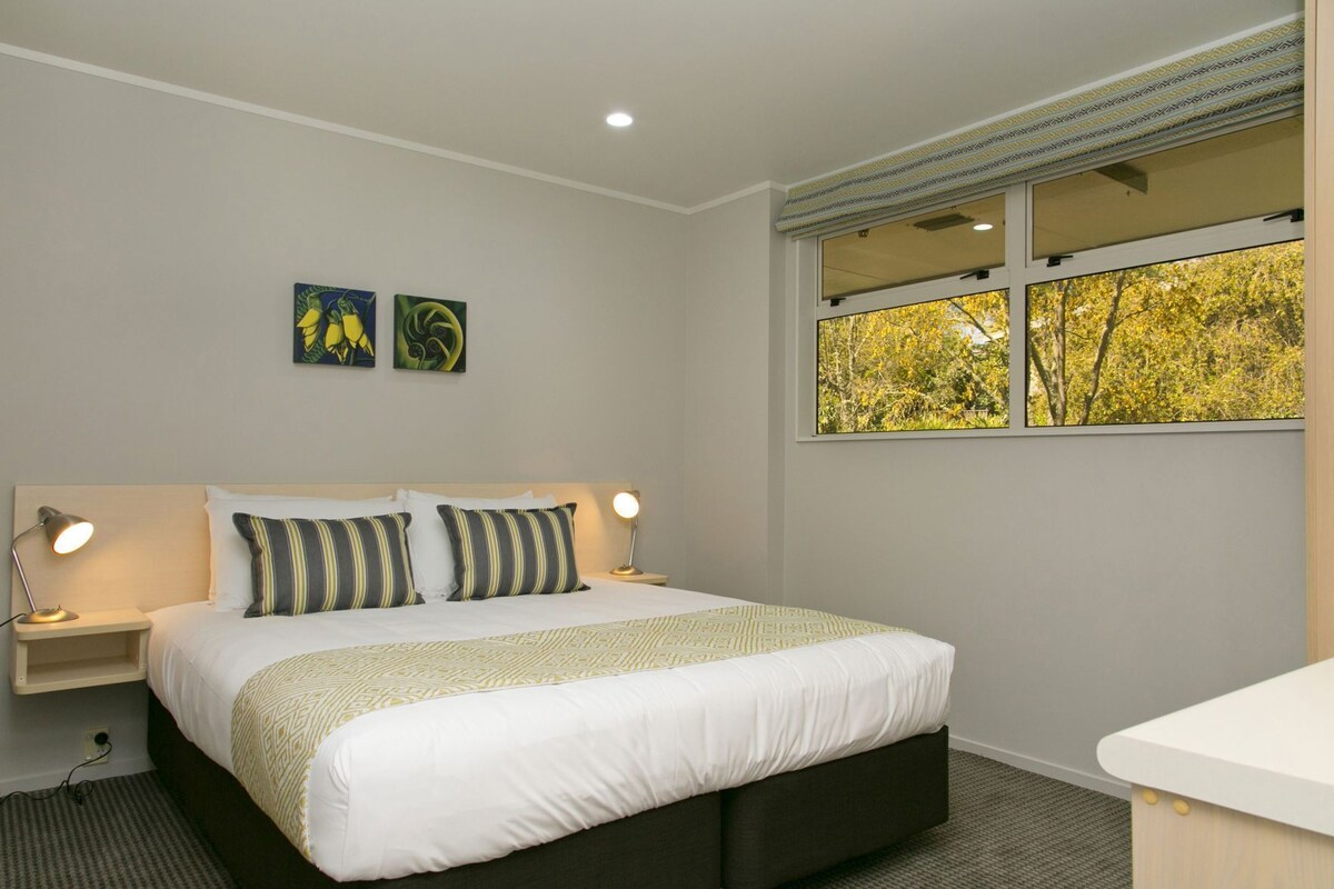 Un dormitorio con una cama, dos lámparas de noche, una mesita de noche, un ventanal con vistas a árboles y dos obras de arte colgadas en la pared.