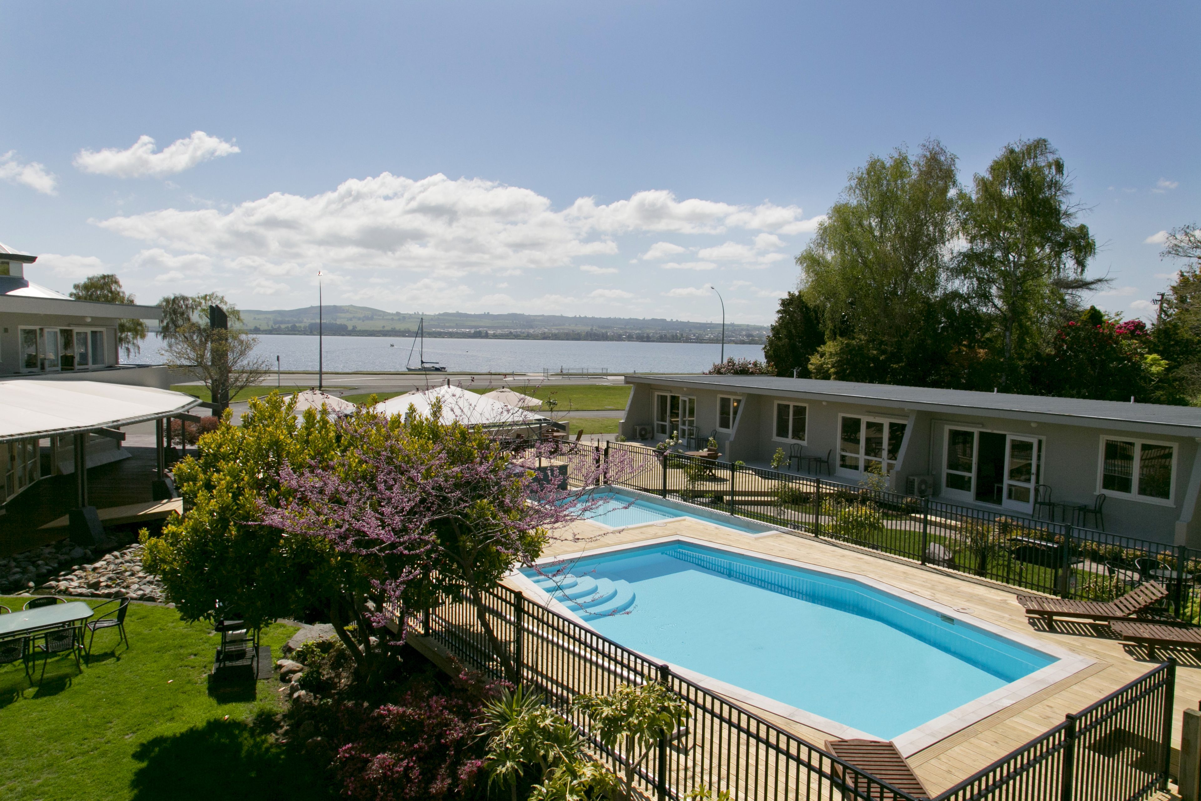 A swimming pool with a view of a lake and mountains, surrounded by a fenced area with lounge chairs.