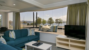 Una sala de estar con sofá, mesa de centro, televisor sobre un mueble bajo y un amplio ventanal con vistas a un lago y árboles.