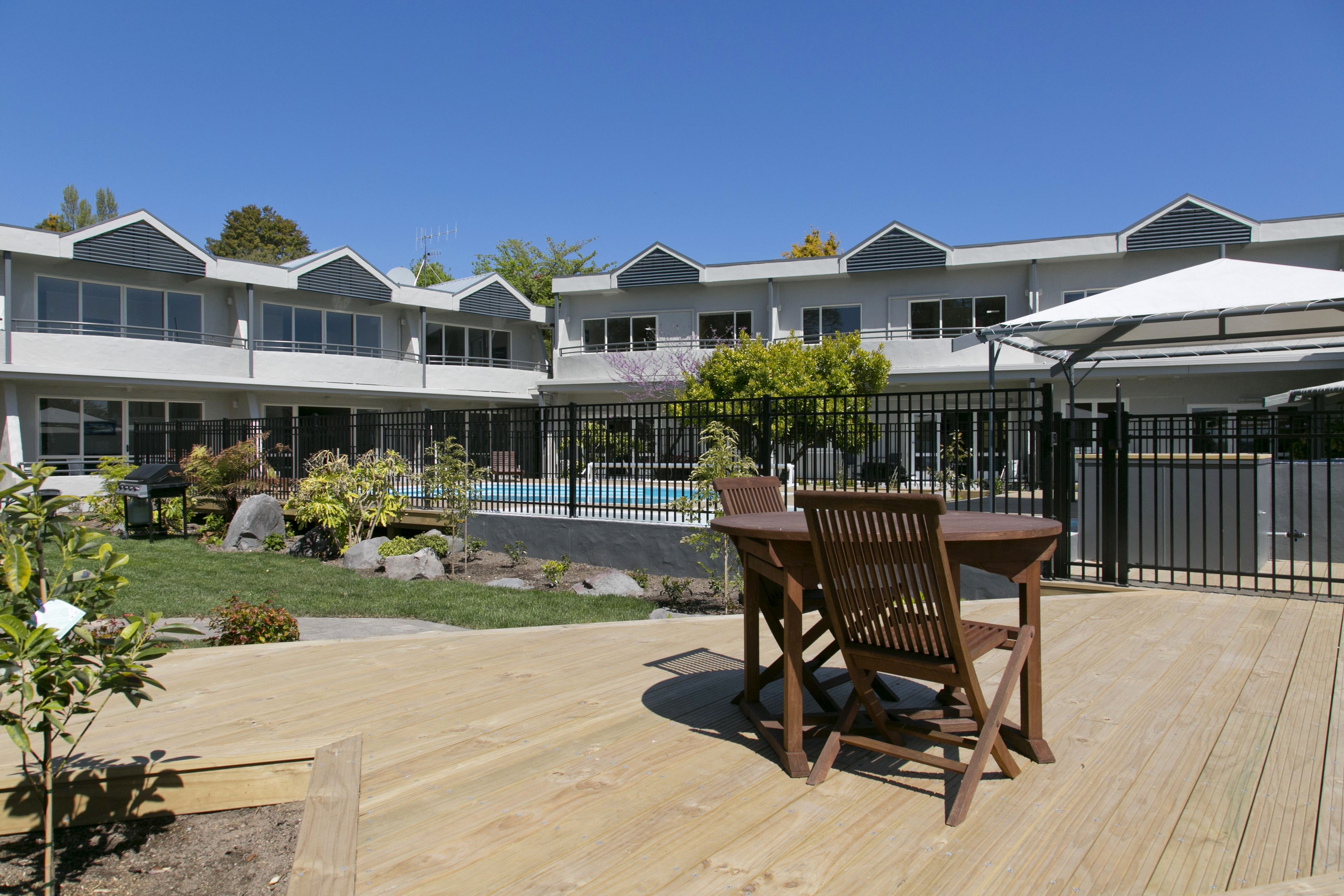 A wooden deck with a table and chairs, a swimming pool, and a building with multiple windows and balconies.