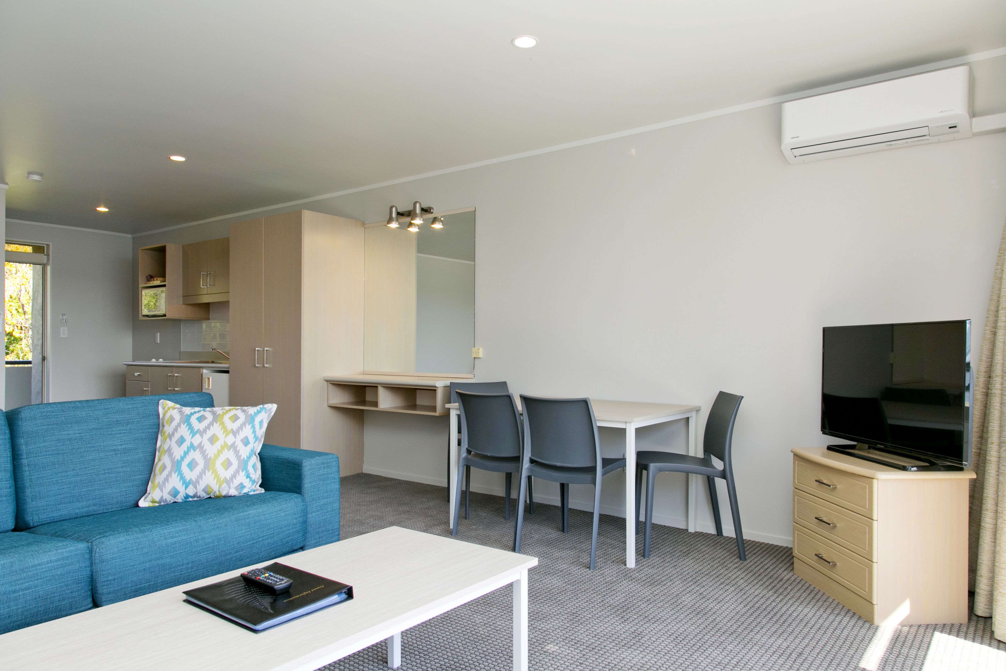 A modern hotel room with a sofa, a dining table with chairs, a TV, and a kitchenette.