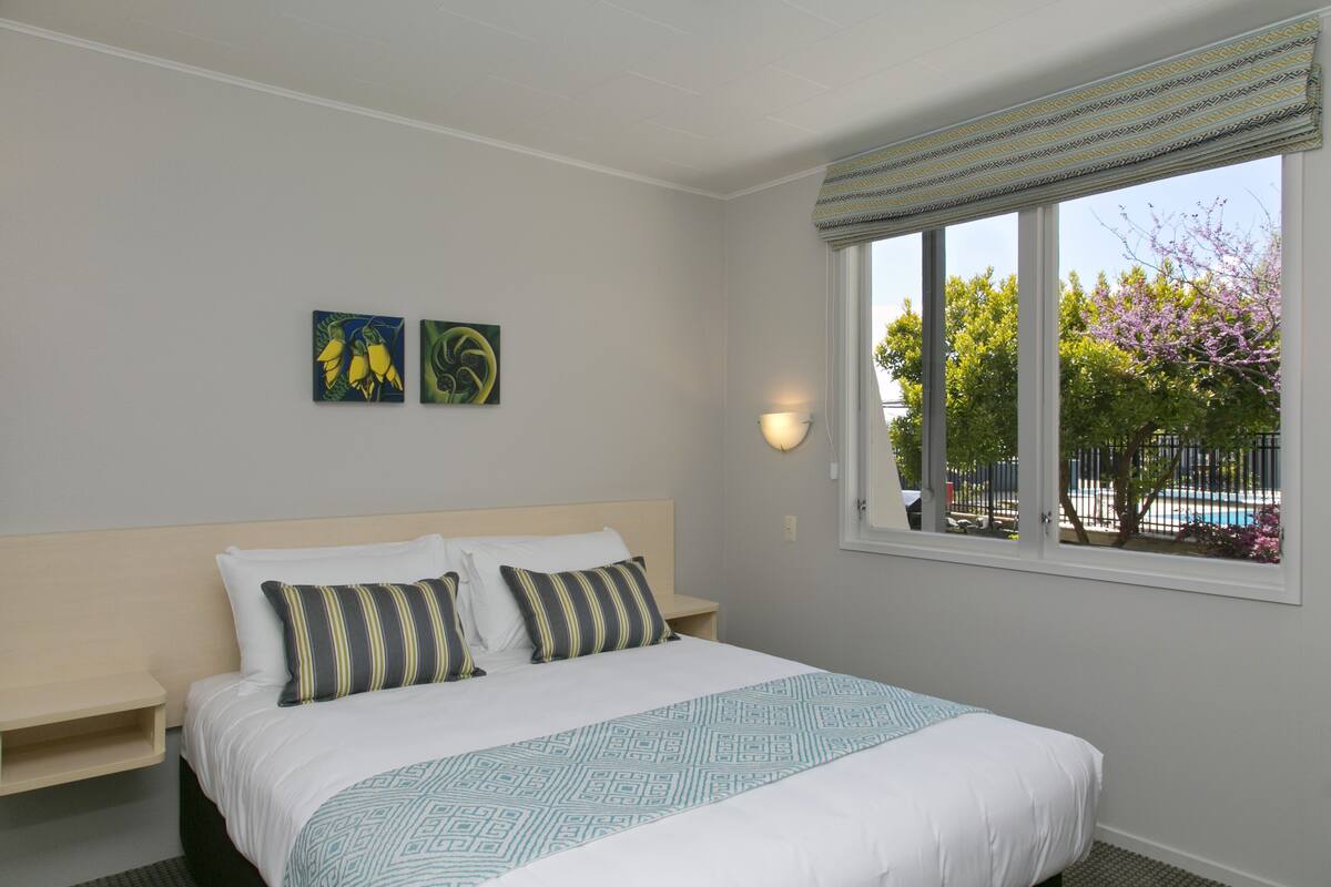 Une chambre d’hôtel avec un lit, deux oreillers, une table de chevet, une lampe fixée au mur et une fenêtre donnant sur des arbres et une piscine.