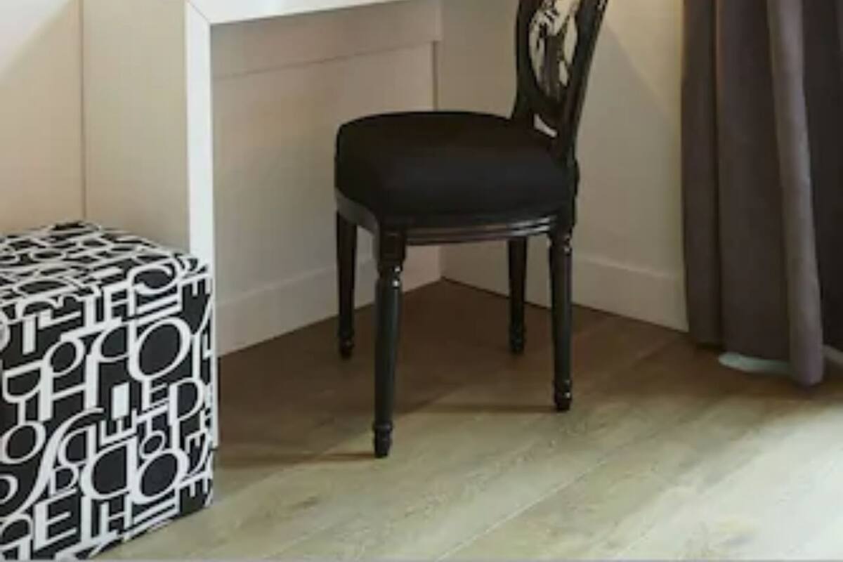 A room with a white desk, a black chair, a television mounted on the wall, a lamp on the desk, and a patterned ottoman.