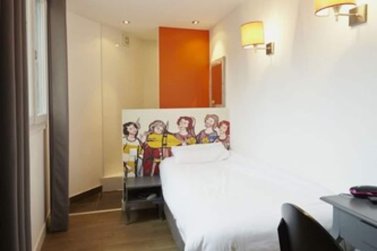 A hotel room with a bed, a desk, and a chair. There is a painting on the wall and a lamp on the desk.