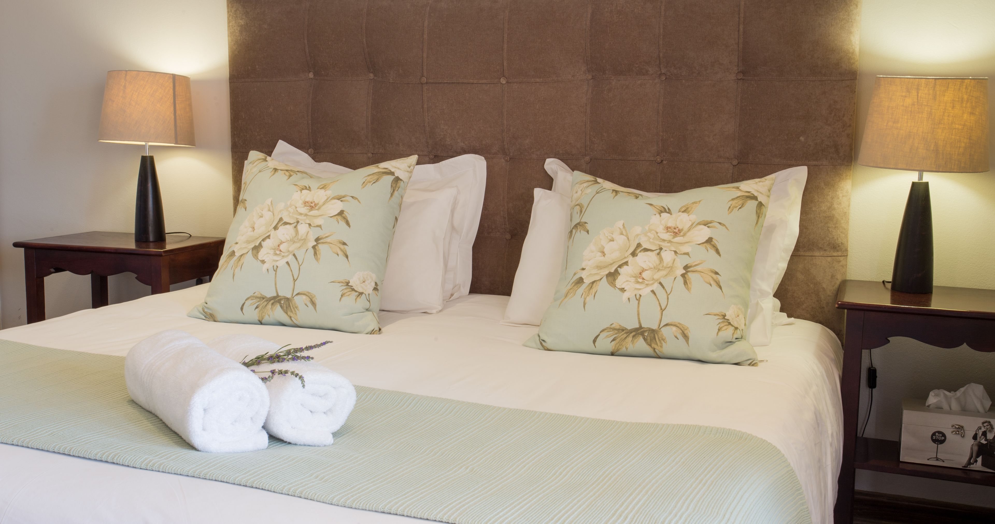 Una cama bien hecha con almohadillas florales, una toalla enrollada y dos lámparas de noche sobre mesitas de madera.