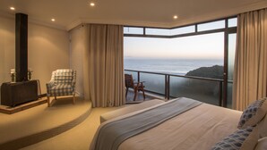 A bedroom with a large bed, a chair, a small table, and a view of the ocean. - Super Tubes Guesthouse (Jeffreys Bay)