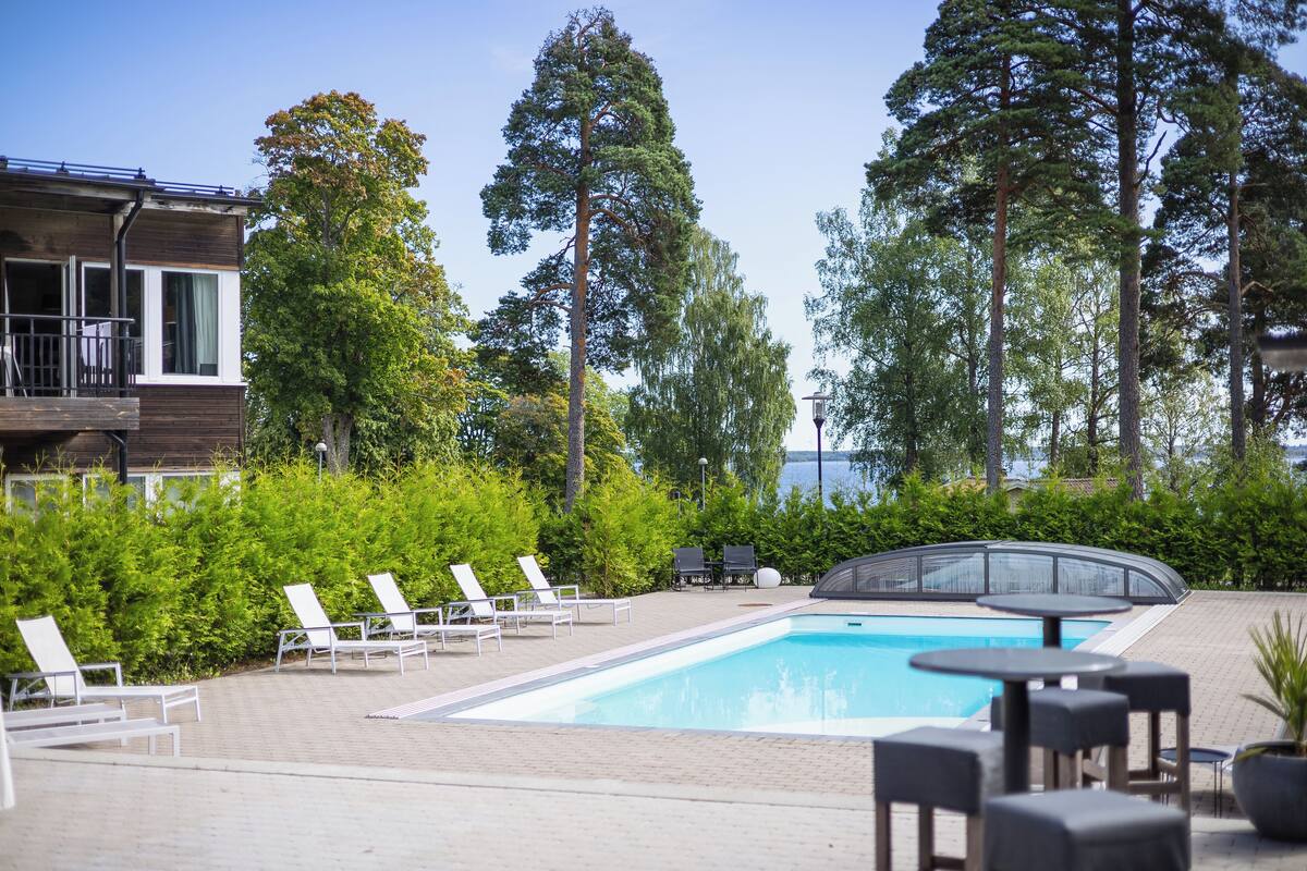 Uima-allasalue, jossa on aurinkotuoleja ja pöytiä, ja jota ympäröivät korkeat puut sekä parvekkeellinen rakennus.