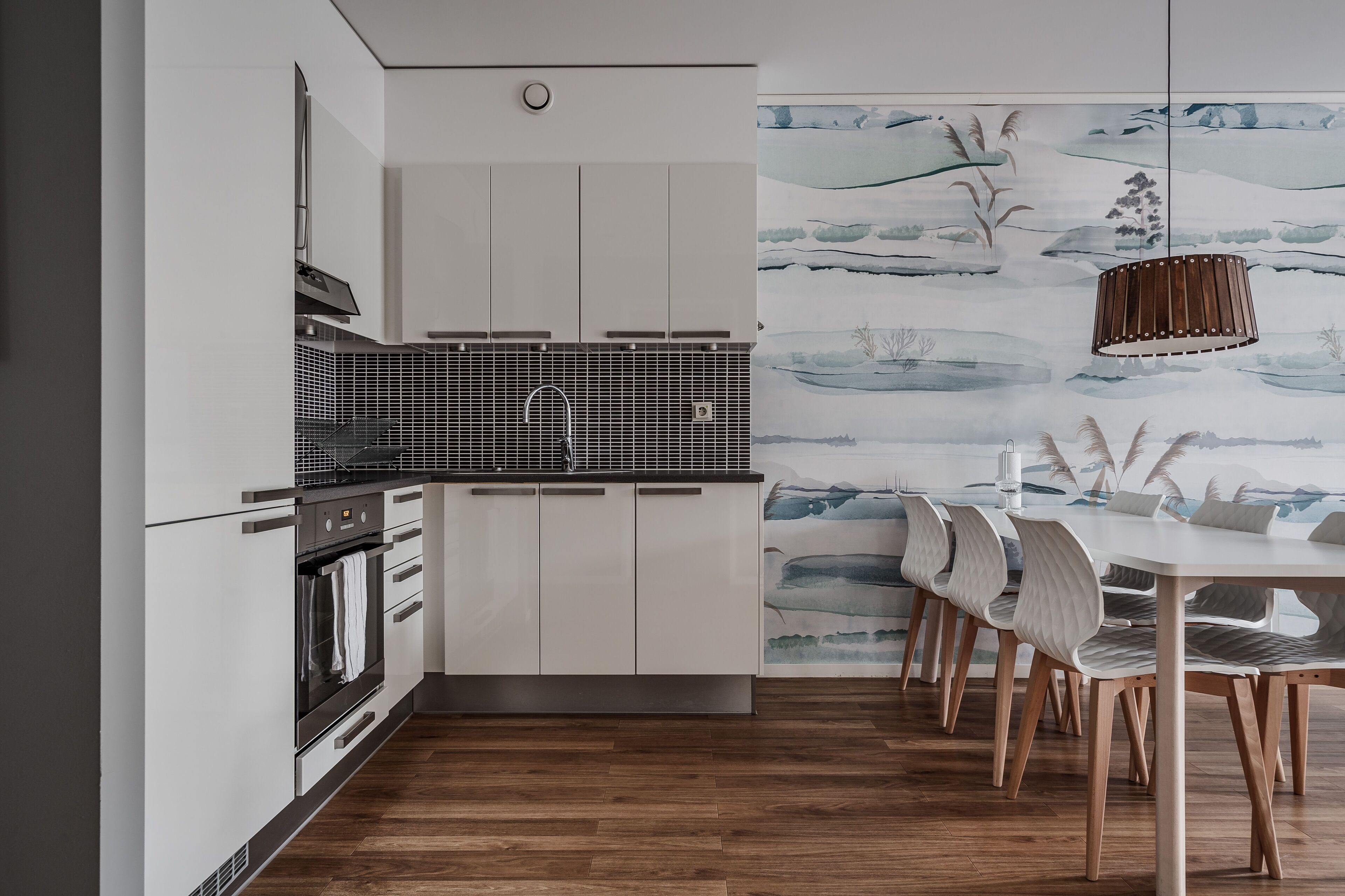 Una cocina moderna con armarios blancos, un protector contra salpicaduras negro y un comedor con sillas de madera y una mesa blanca.