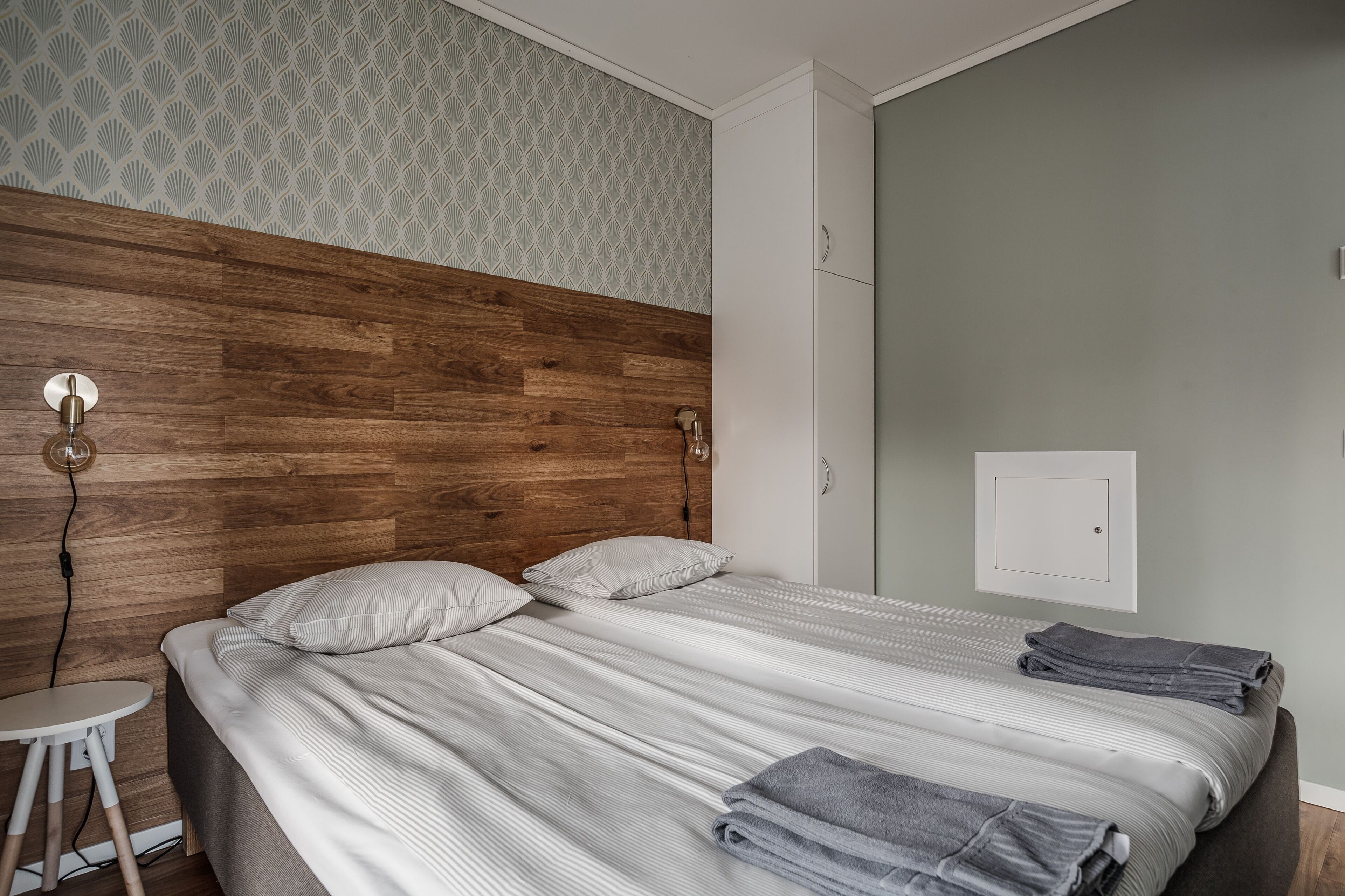 A neatly made bed with a striped bedspread and two pillows, a wooden headboard, a small white table, and a wall-mounted light fixture.