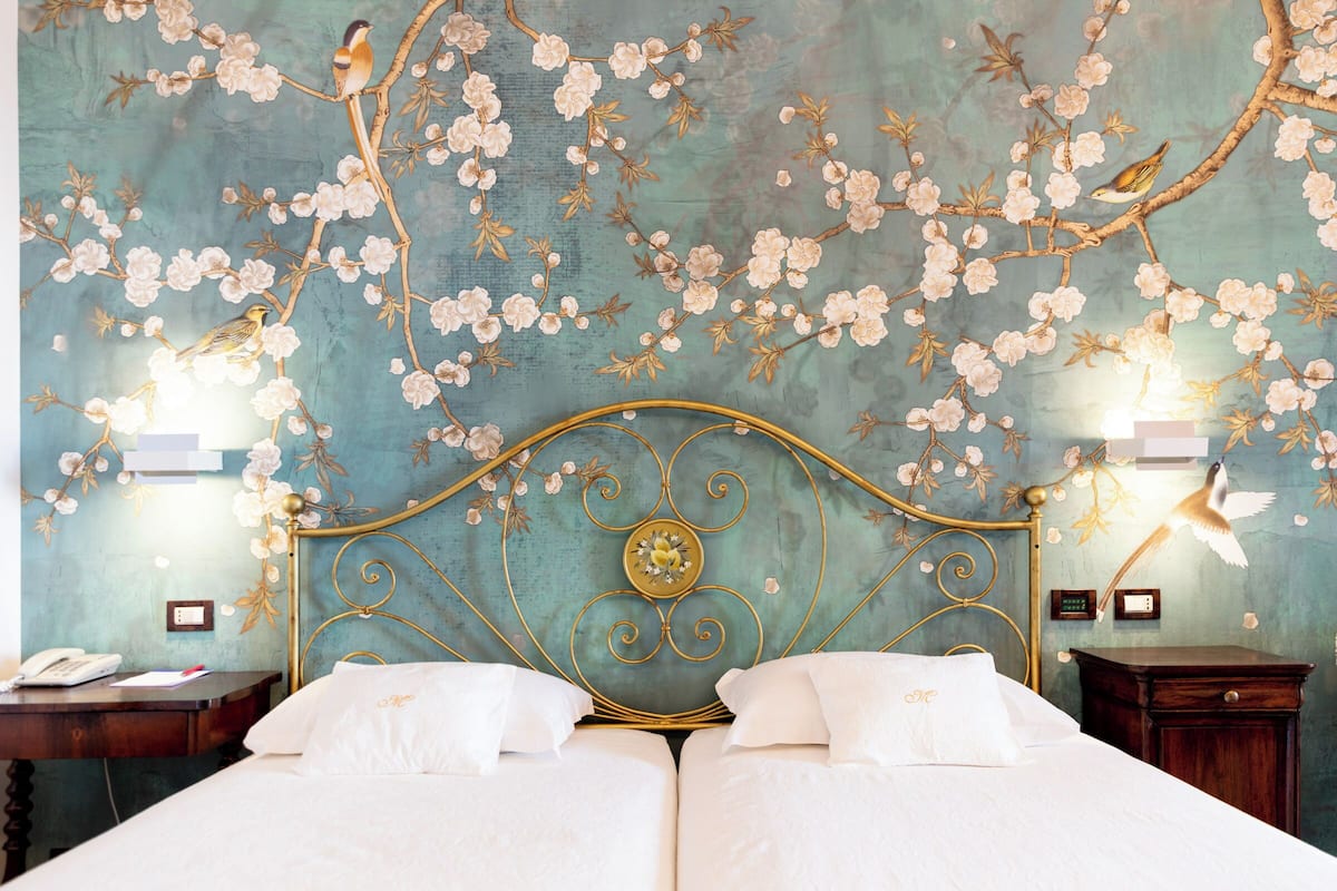 Un lit double agrémenté d’une tête de lit décorative, une table de chevet avec une lampe et un mur recouvert d’un papier peint à motifs floraux.