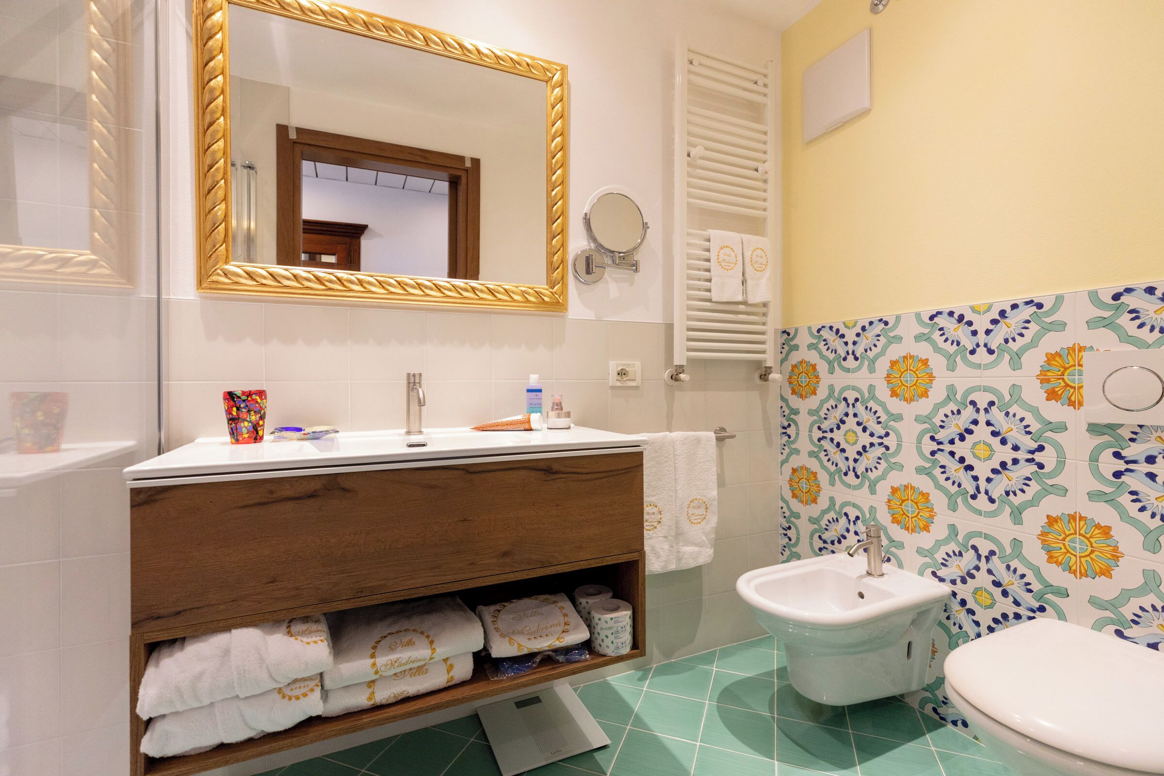 Un baño con tocador de madera, lavamanos, inodoro, espejo y baldosas estampadas.