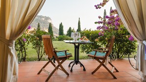 Eine Terrasse im Freien mit zwei Holzstühlen und einem Tisch, umgeben von Vorhängen und Blick auf einen Garten mit Blumen und Bäumen.