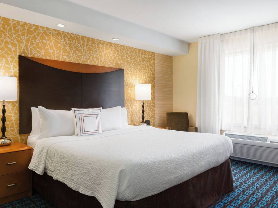 A hotel room with a large bed, two bedside lamps, a nightstand, a chair, and an air conditioning unit.