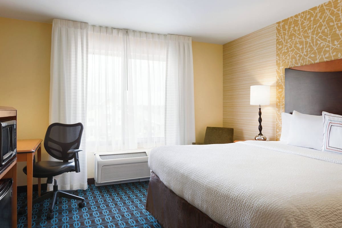 A hotel room with a bed, a desk with a chair, a lamp, and a window with curtains.