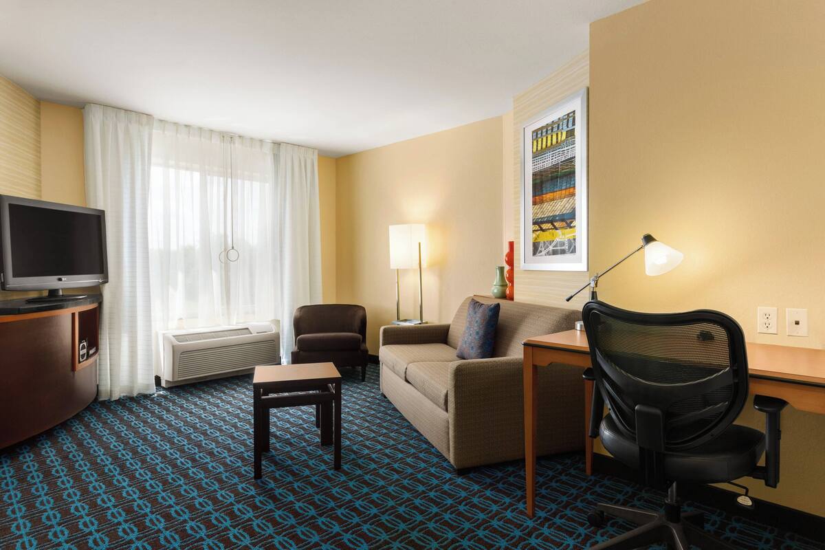 A hotel room with a television, a sofa, a desk, a chair, and a lamp.