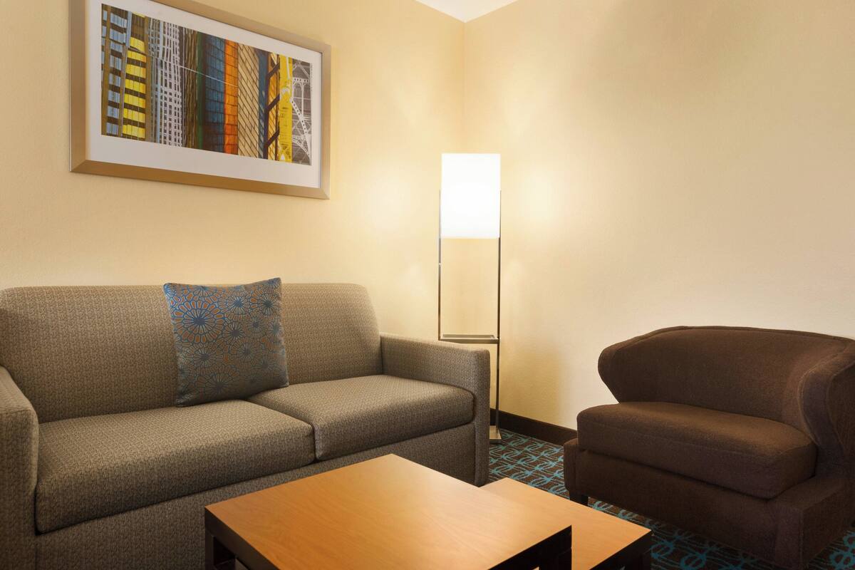 A hotel room with a grey sofa, a brown armchair, a wooden coffee table, and a floor lamp.