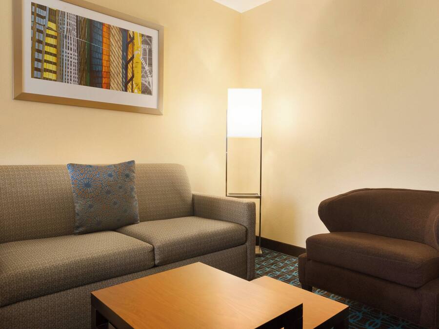Een hotelkamer met een grijze bank, een bruine fauteuil, een houten salontafel en een vloerlamp.