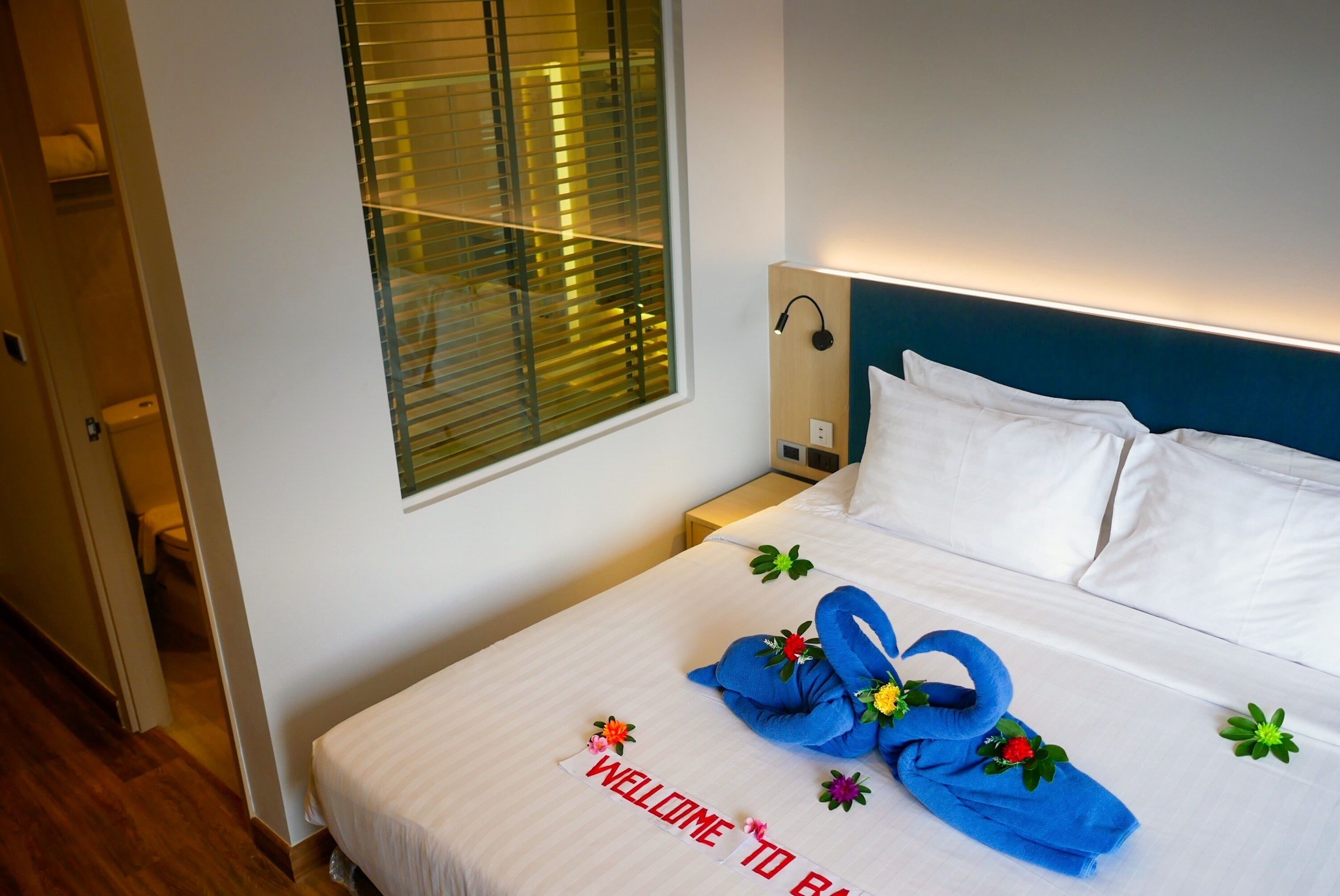 A hotel room with a neatly made bed, a bedside table with a lamp, and a window with blinds.