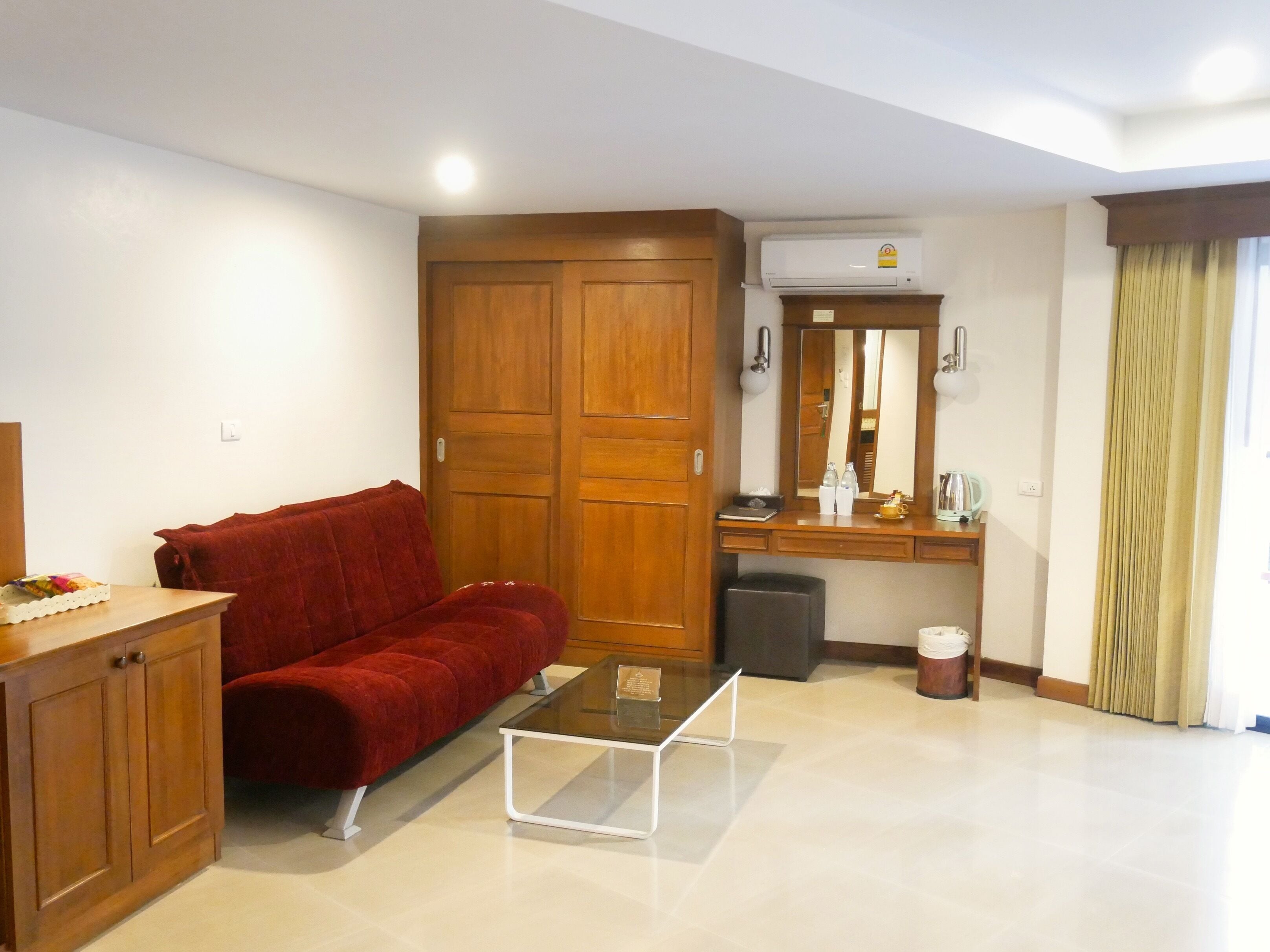 Un ambiente compatto con un divano rosso, un tavolino con piano in vetro, un armadio in legno, uno specchio e un piccolo cestino.