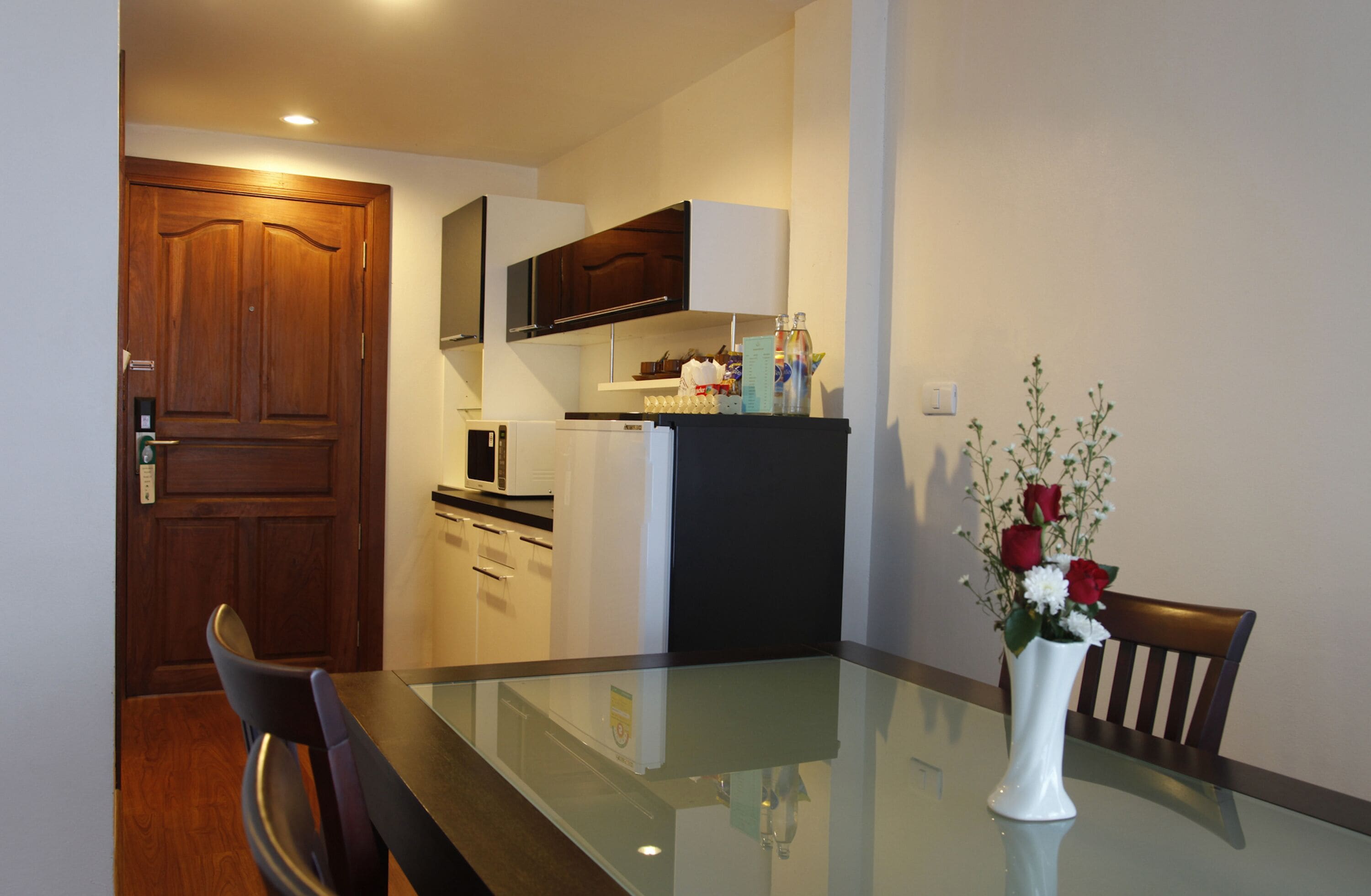 A compact kitchen with a microwave, refrigerator, and wooden cabinets. A dining table with chairs is in the foreground, and a vase with flowers is on the table.