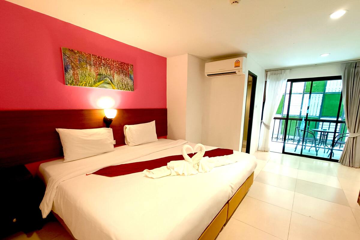 A hotel room with a bed, a painting on the wall, an air conditioning unit, and a view of the outside.