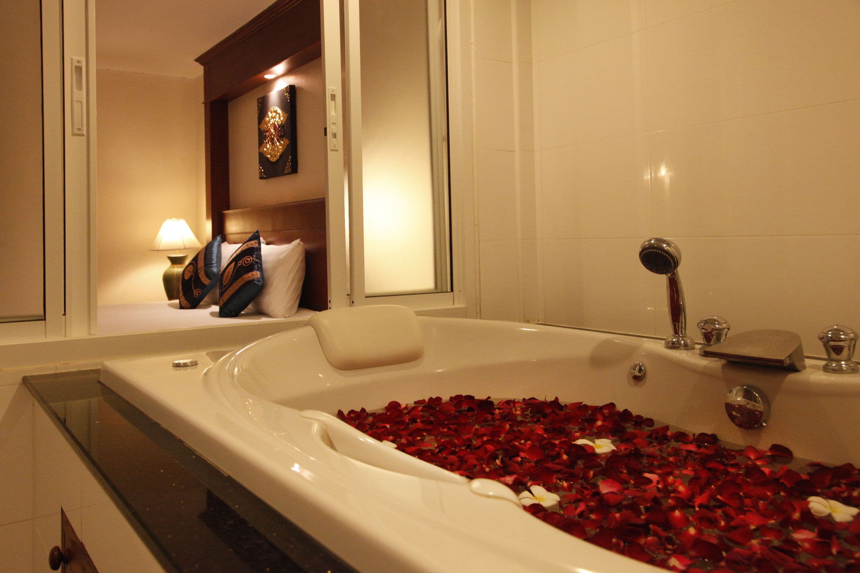 A bathtub filled with rose petals and a mirror reflecting a room with a bed and a nightstand.