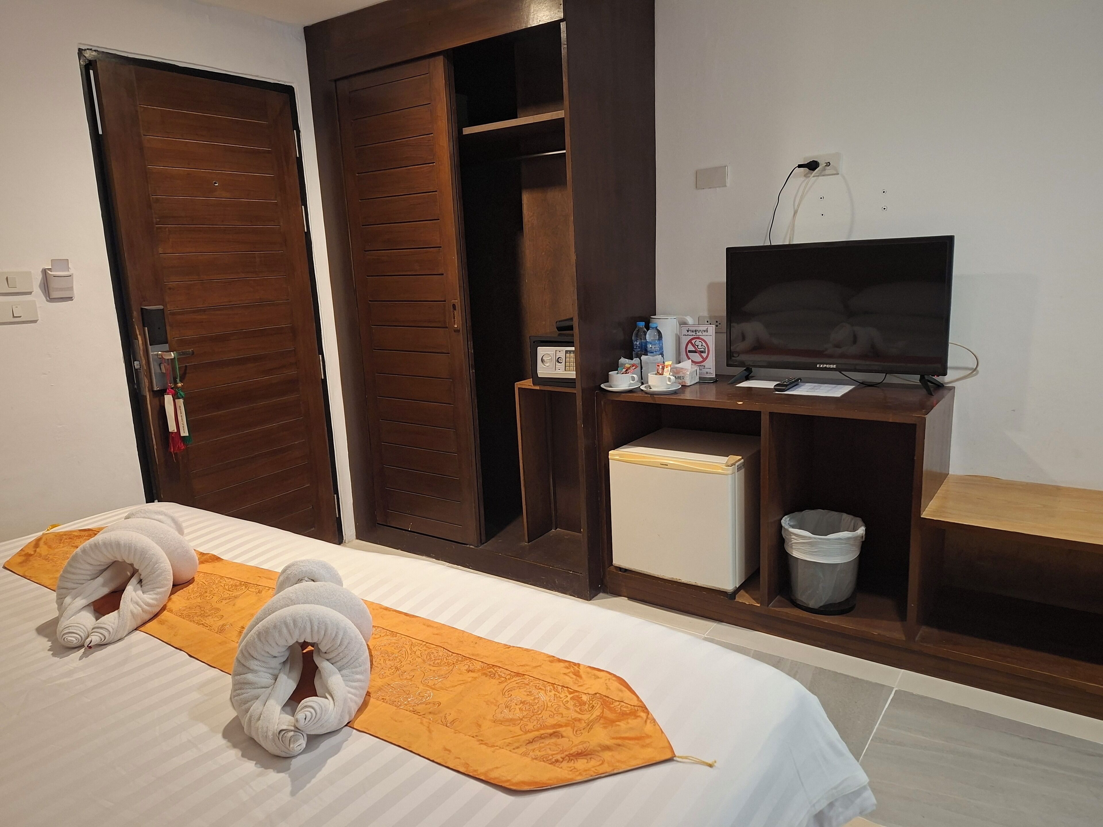 A hotel room with a bed, two rolled-up towels, a television, a mini-fridge, a trash bin, and a wooden wardrobe.