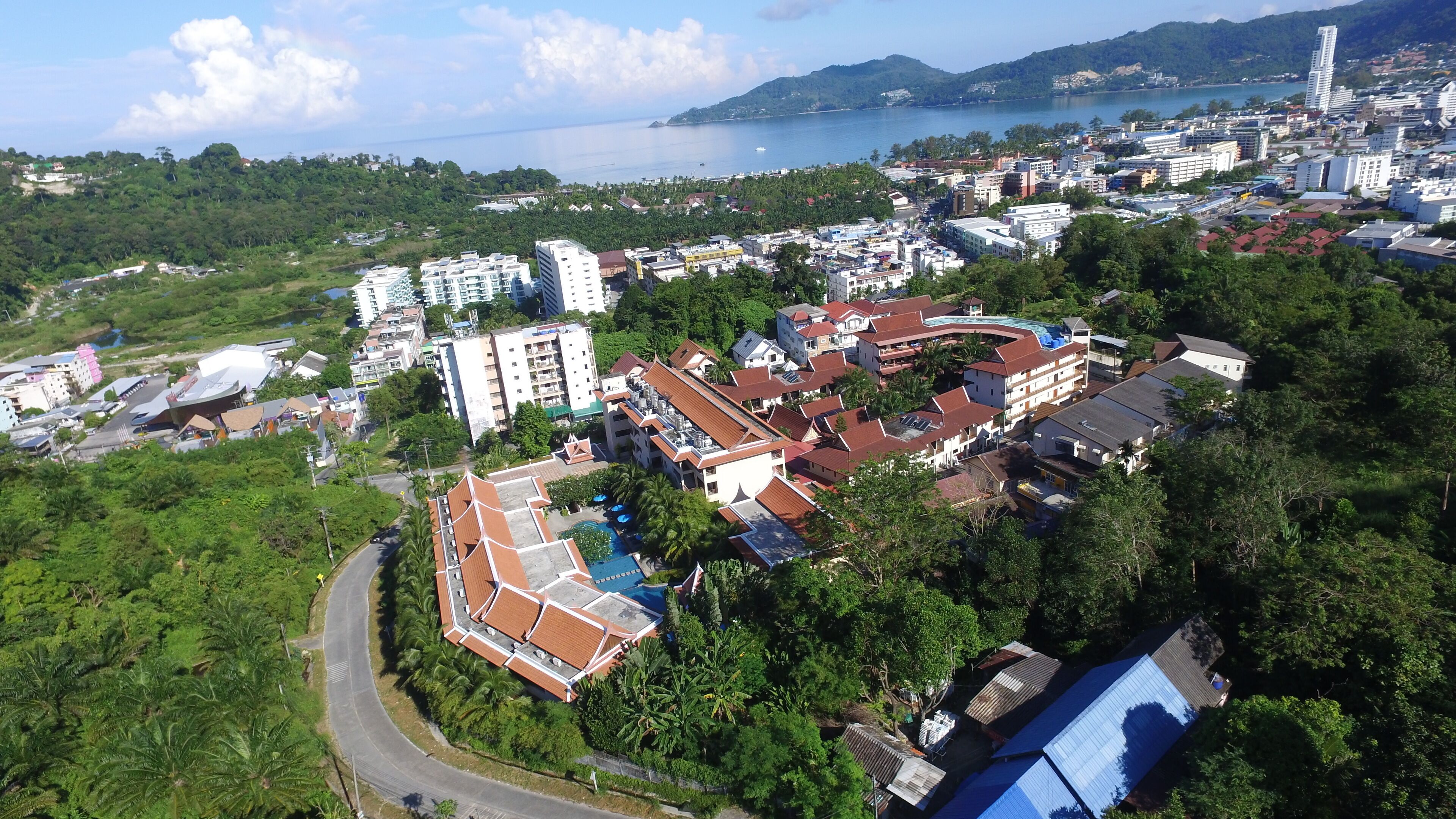 An aerial view of a coastal city with a mix of residential and commercial buildings, a prominent hotel with a swimming pool, and a view of the sea.