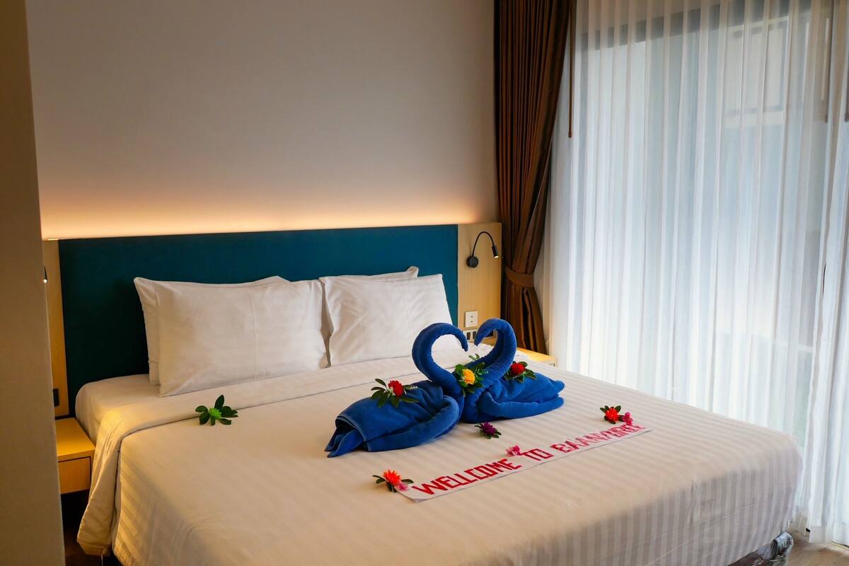 A hotel room with a neatly made bed, a blue headboard, and a welcome sign on the bed.
